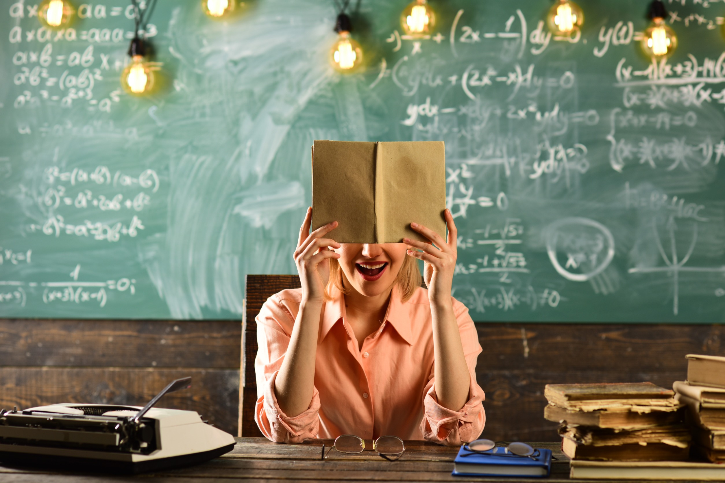 educadora con libro en la cara y pizarra con bombillas encendidas de fondo